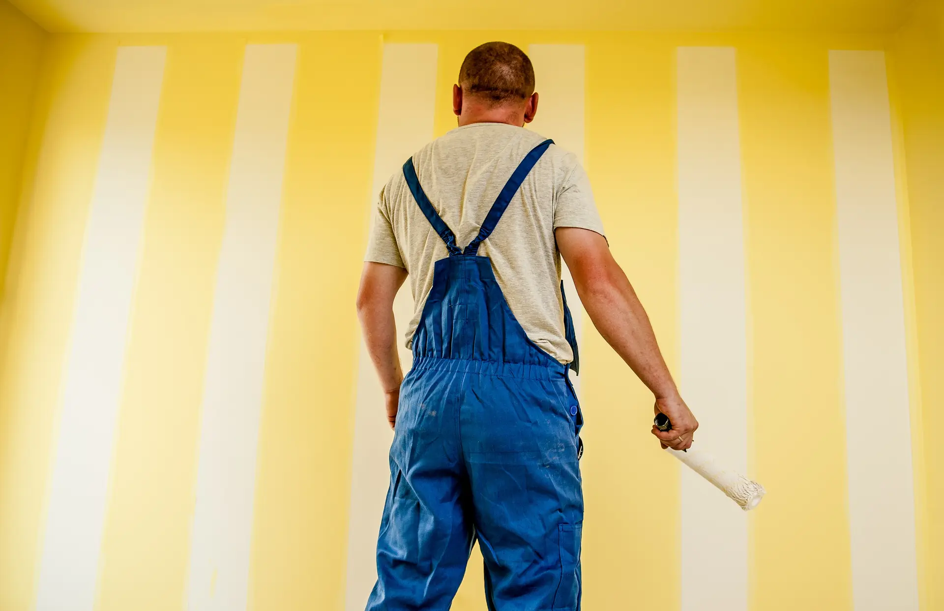 Un compagnon en bleu de travail face à un mur jaune en cours de finition